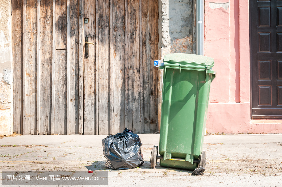 一个绿色的垃圾桶和黑色的塑料垃圾袋在城市的街道上,等待倾卸卡车收集在房子前面
